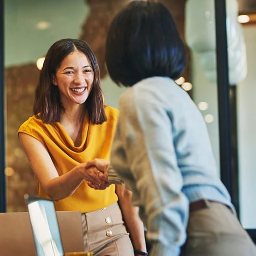 Two people shake hands in an office, one in a yellow top and the other in a gray sweater, suggesting a professional meeting.