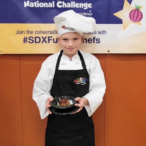 kid in a white chef's hat and black apron with the 'Future Chefs National Challenge' logo, presenting a plated dish in front of a banner featuring the 'National Challenge' and 'Sodexo' logos