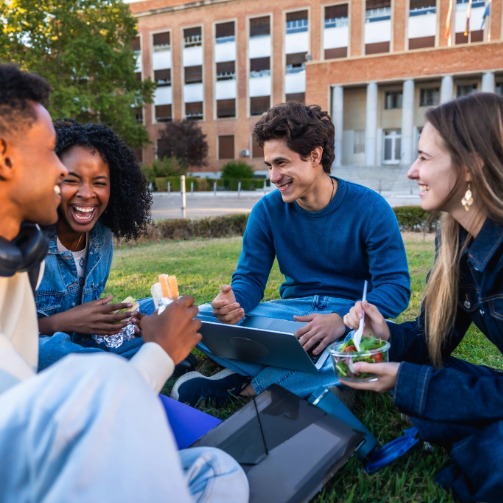 Friends enjoying food and laughing on a college campus lawn.