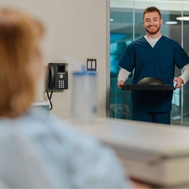 Smiling healthcare staff member delivering a meal tray to a patient in a hospital room. 