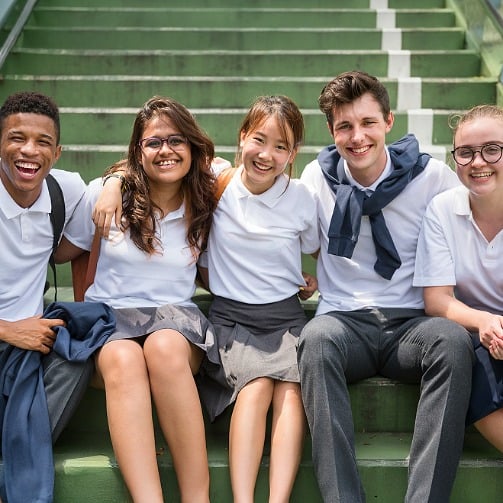 Students in uniform sit on outdoor steps, smiling brightly with their arms around each other.