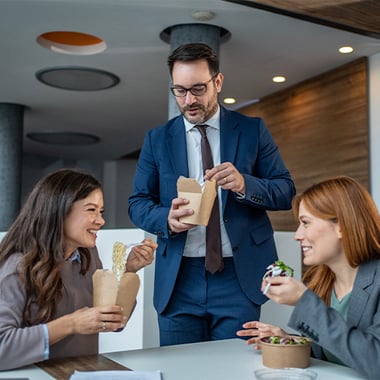 Three coworkers share a casual meal around a table in a modern office.