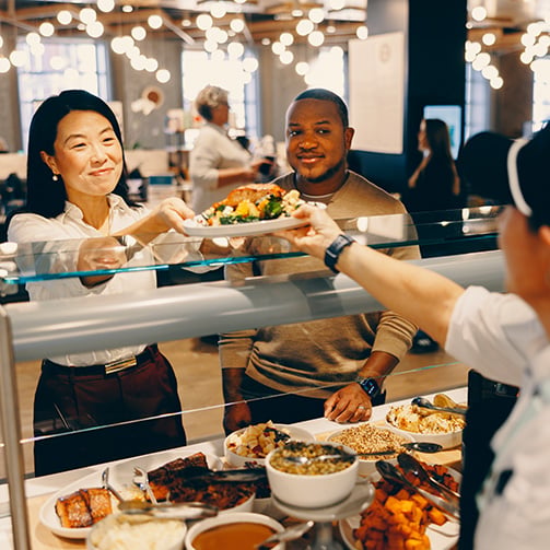 Customer receiving a plated meal at a cafeteria counter with servers and various dishes visible in background.