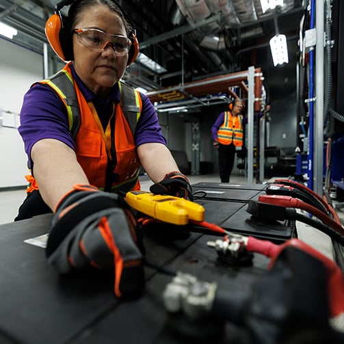 Technician in safety gear testing large batteries with a multimeter in an industrial data center.