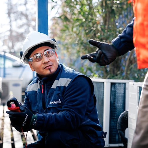 Worker wearing a hard hat and safety glasses examining equipment outdoors, holding testing tools while another gloved hand gestures nearby in an industrial setting.