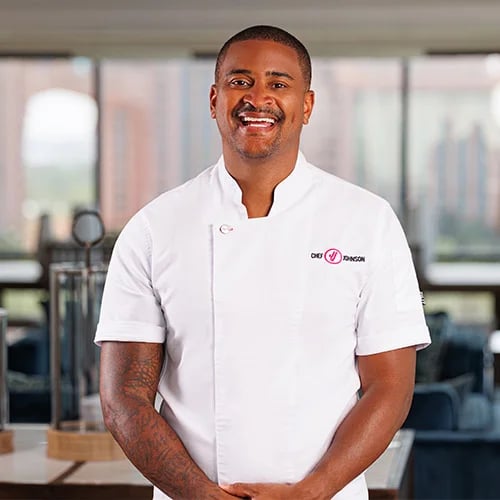 A man wearing a white chef's coat stands confidently in a kitchen setting.