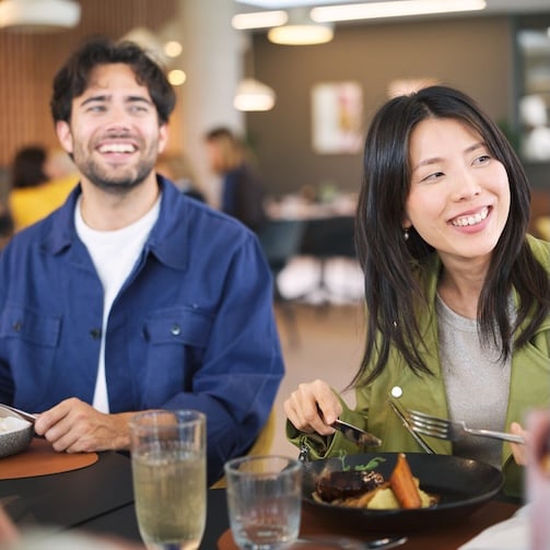 Two diners seated at a restaurant table eating meals with drinks in a modern café setting.