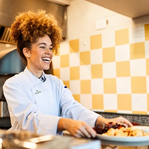  A smiling chef in a kitchen handing over a plated dish.