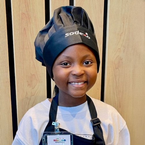 Kid wearing a black chef's hat with the 'Sodexo' logo, a white shirt, and a black apron, standing in front of vertical wooden panels, with a name badge attached to the apron.
