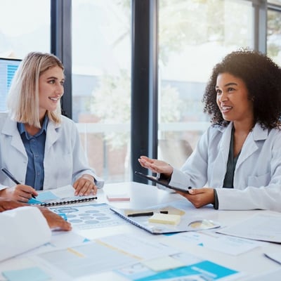 Two dietitians sit at a table reviewing research notes, with one holding a tablet and data displayed on a screen behind them.