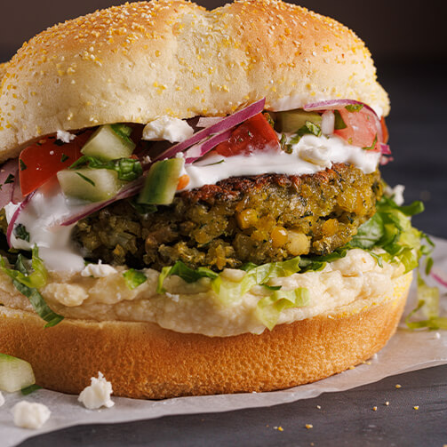 Image of a close-up sandwich with a golden bun and a vegetable patty. It's layered with diced cucumbers, tomatoes, red onions, crumbled feta cheese, a drizzle of creamy white sauce (likely tzatziki), and shredded lettuce, all resting on parchment paper with a few scattered bits of ingredients