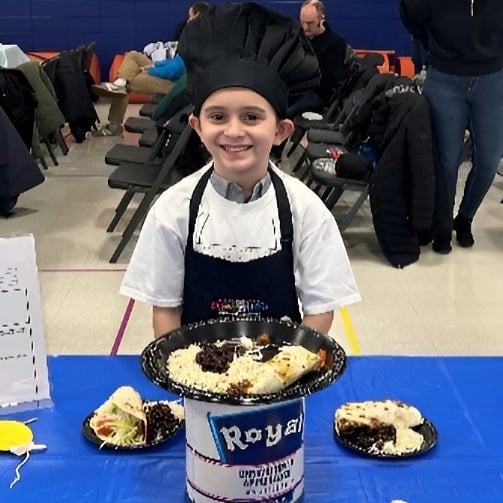 Child in chef hat and apron standing at table with plated dishes at a school cooking event.
