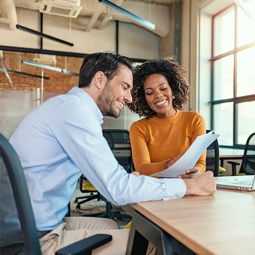 A man and woman smiling and sit at a table, focused on a laptop in front of them.