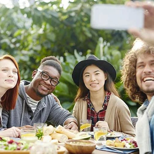 Group of people sitting at a table outdoors, enjoying a meal together while one person takes a photo with a smartphone.
