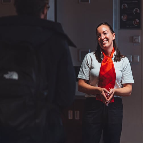 Person in gray shirt with red scarf stands indoors speaking to someone wearing a backpack.