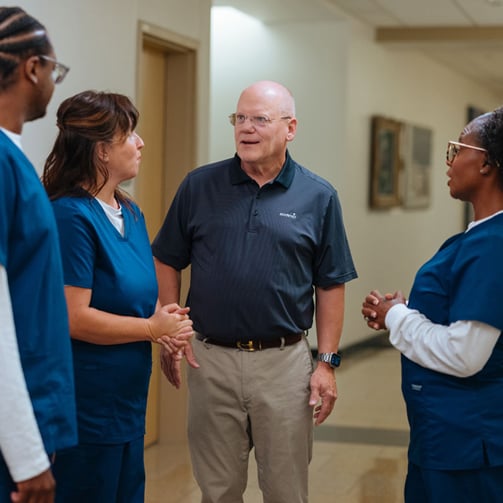 Person in a polo shirt speaking with three healthcare staff members in a hospital hallway.