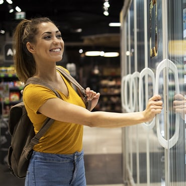 Use alt text: A female student, wearing a backpack, smiles while browsing the refrigerated section of a convenience store.