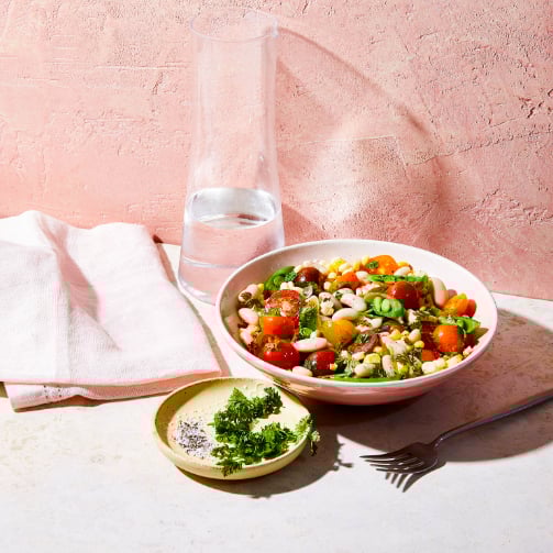 A colorful bowl of vegetables, beans and herbs sits beside a clear pitcher of water in a warm, inviting setting.