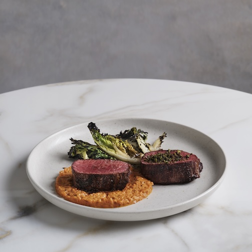 Sliced steak medallions with greens and puree on a white plate set on a marble table.