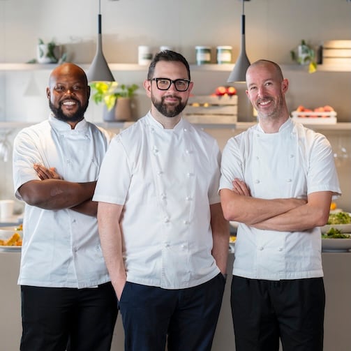 Three chefs in white uniforms standing with arms crossed in a bright modern kitchen.