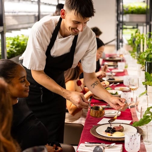 Server plating a dish at a long dining table with seated guests in a bright, modern event setting.