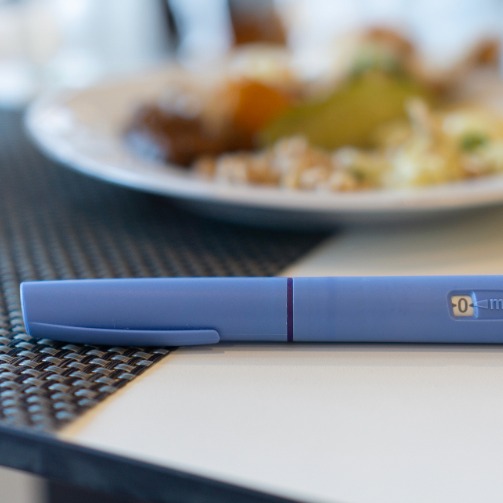 Close-up of a blue injection pen placed on a table next to a meal, with a plate of food blurred in the background, suggesting a connection between medication and dietary context.