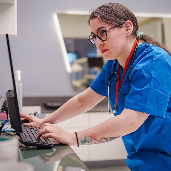 Healthcare professional in blue scrubs using a desktop computer at a clinical workstation, with a stethoscope, identification badge, and keyboard visible in a modern healthcare environment.
