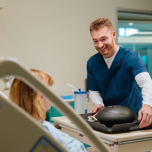 Healthcare worker in blue scrubs placing a covered meal tray on a bedside table for a patient in a hospital room, with medical furnishings and care equipment visible nearby.