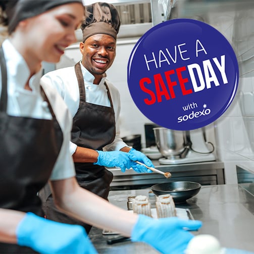 Food service workers preparing meals in a commercial kitchen beside a “Have a Safe Day” safety message.