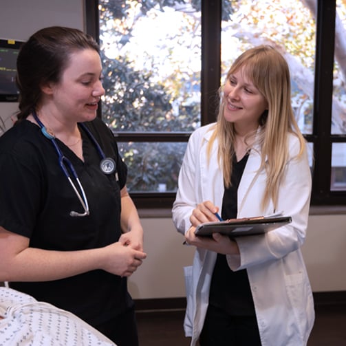 Copilot said:  Healthcare professional in medical scrubs speaking with a colleague in a white lab coat holding a tablet, standing beside a patient bed in a bright clinical setting with large windows.