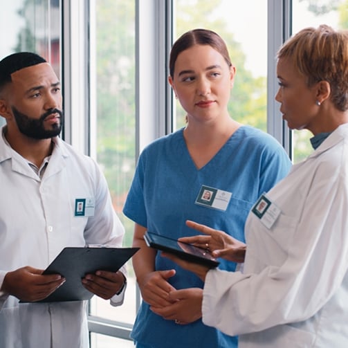 Copilot said:  Three healthcare professionals standing together in a bright clinical setting, reviewing information on tablets and clipboards near large windows, wearing medical scrubs and lab coats with identification badges.