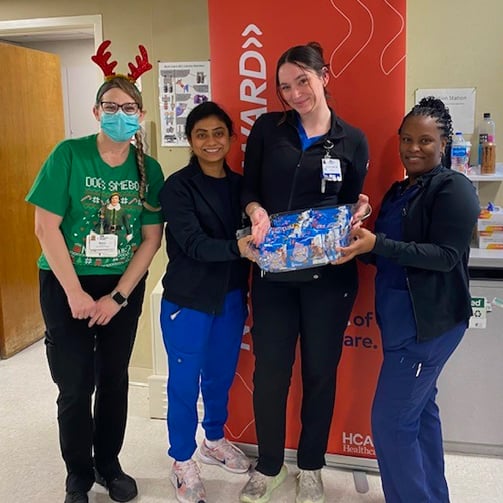 Four healthcare staff stand indoors holding a basket, one wearing reindeer antlers and a face mask.