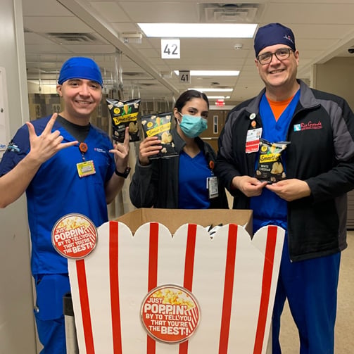 Three healthcare workers hold snack bags behind a large popcorn box display in a hospital hallway.
