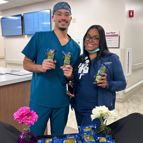 Two healthcare workers holding packaged snacks, standing behind a table with flowers and more snack items.