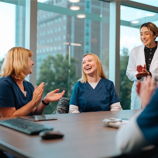 Healthcare staff seated around a conference table in a glass-walled meeting room, participating in a team discussion, with laptops and notebooks visible.