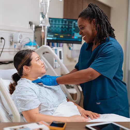 Healthcare worker adjusting bedding for a patient resting in a hospital bed, with medical monitors and equipment visible in a clean, well-lit clinical room.