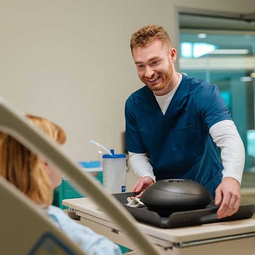 Healthcare worker placing a meal tray with covered dishes and a drink on a bedside table for a patient resting in a hospital room with clinical equipment nearby.