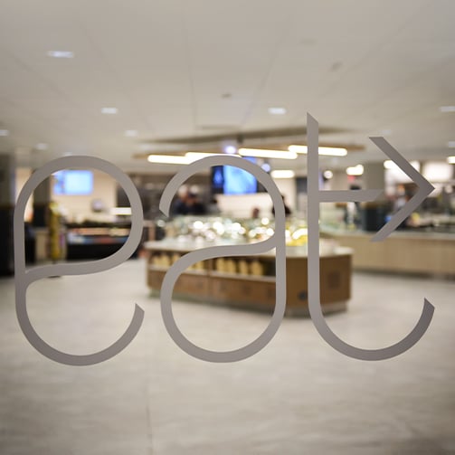 Frosted glass sign reading “eat” with an arrow, looking into a modern cafeteria with food stations beyond.