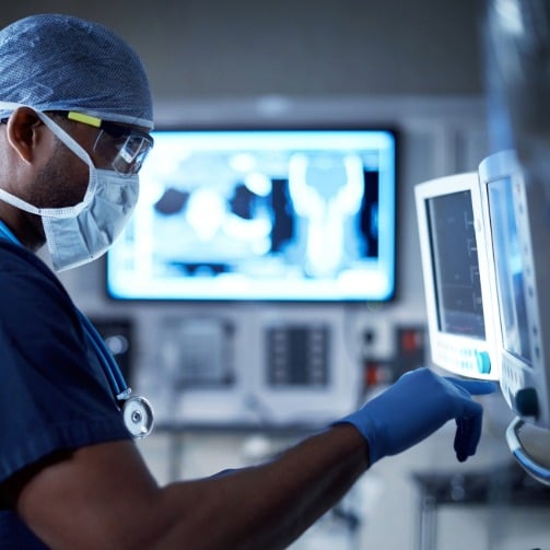 Doctor in surgical attire and mask monitoring medical equipment in a hospital setting.