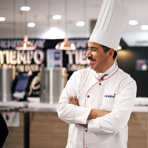 A chef wearing a white hat stands in a kitchen, surrounded by cooking equipment and ingredients.