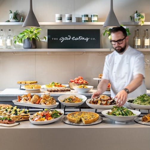 Chef arranging colorful buffet dishes on a counter beneath “The Good Eating Co.” sign in a modern kitchen setting.