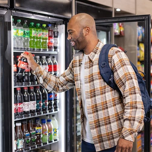 Person in plaid shirt and backpack grabs a soda from a fridge stocked with assorted beverages.
