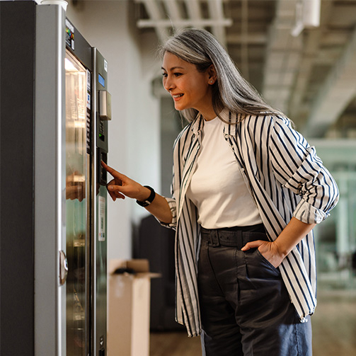 Person with long gray hair, wearing a striped shirt and dark pants, uses a vending machine in a modern indoor space.