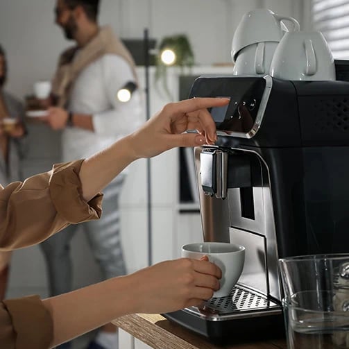 Person uses espresso machine to pour coffee while two others chat in blurred background.