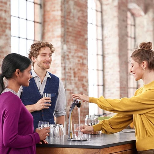 Three people at a counter hold water glasses while one fills a glass from a water dispenser.