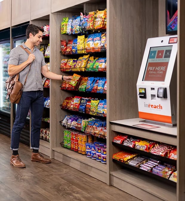 Person choosing snacks from a shelf beside a self-checkout kiosk in a convenience store.