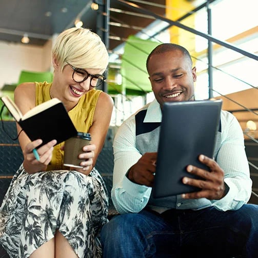 Two people sit on stairs, one holding a book and coffee, the other a tablet, engaged in reading in a modern setting.