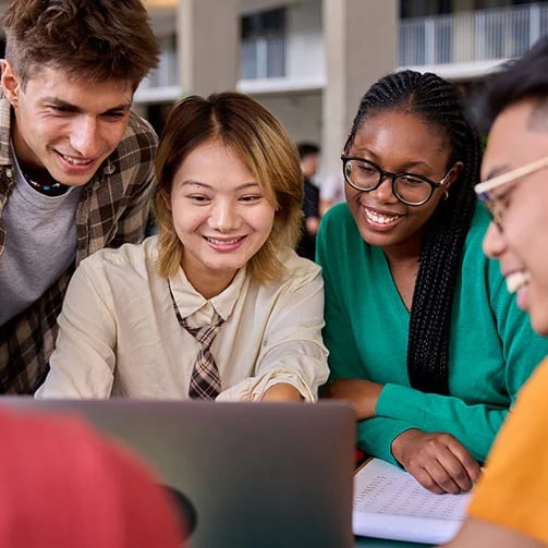 A group of students gathered around a laptop, engaged in discussion and looking at the screen together.