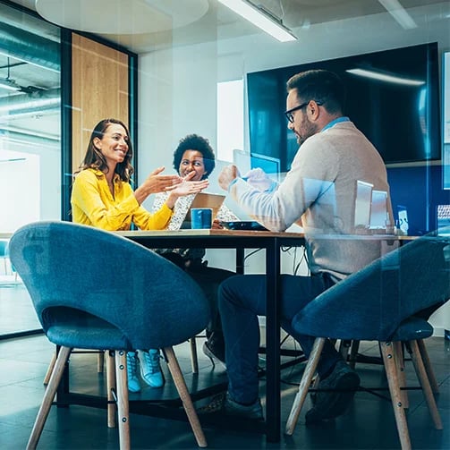 Three people have a meeting in a modern office with glass walls, sitting around a table with laptops and coffee cups.