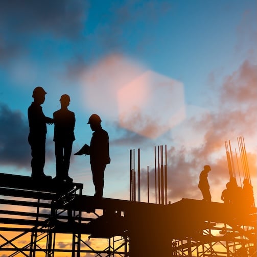 Silhouetted construction workers on scaffolding at sunset with exposed steel supports in the background.